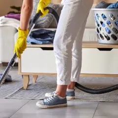 Persona con guantes amarillos usa una aspiradora para limpiar la alfombra. Hay una cesta de ropa sucia y un sofá en el fondo.