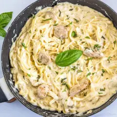 Un cremoso plato de pasta alfredo con trozos de pollo y hojas de albahaca fresca en una sartén.