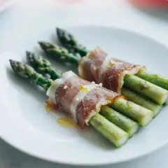Zielone szparagi owinięte w cienkie plastry szynki parmeńskiej, ułożone na białym talerzu.