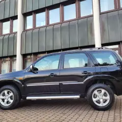Czarny Mazda Tribute stoi na brukowanym podjeździe przed nowoczesnym budynkiem.