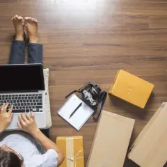 Osoba pracuje na laptopie otoczona paczkami, jakby przygotowywała się do wysyłki. To idealny scenariusz dla kuriera InPost.