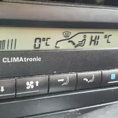 Panel klimatyzacji w samochodzie. Wyświetla 0°C, ikonę nawiewu na szybę i pasażerów, a także ustawienie "HI".