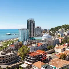 Widok na miasto Durres z plażą w tle. Widać morze, promenadę, budynki mieszkalne i hotelowe oraz wzgórze porośnięte zielenią.