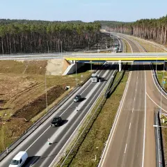 Droga ekspresowa z mostem, samochodami i ciężarówką. W tle las. Trudno ocenić, ile km na drodze ekspresowej.