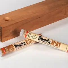 Two tubes of WoodFil Epoxy, a wood repair putty, lie next to a wooden plank with a hole, demonstrating how to use wood filler for quick repairs.