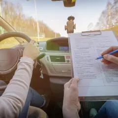 Egzamin na prawo jazdy. Kandydat trzyma kierownicę, a egzaminator zaznacza na liście kontrolnej różne kategorie prawa jazdy.