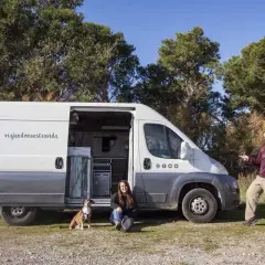 Pareja y perro posan junto a su furgón vivienda, listos para viajar.