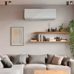 Un aire acondicionado blanco en la pared controla el calor, mostrando 26°C. Debajo, un sofá gris con cojines y estanterías con libros y plantas.