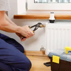 Instalator dokręca kluczem złączkę S do grzejnika, przygotowując go do sezonu grzewczego. Obok stoi skrzynka z narzędziami.