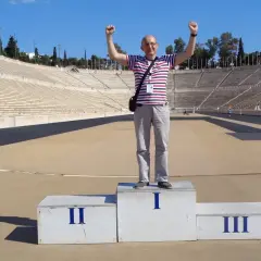 Mężczyzna na podium w historycznym stadionie panatenajskim, z rękami w górze, świętuje zwycięstwo.