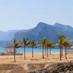 Oman co warto wiedzieć: piaszczysta plaża z palmami, biała altana i turkusowe morze na tle gór. Idealne miejsce na relaks.