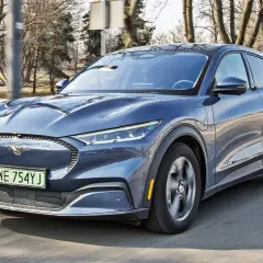 Niebieski Ford Mustang Mach-E jedzie po drodze, z widocznym logo konia na grillu.