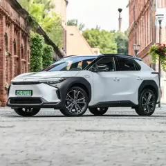 Biały Toyota bz4x na brukowanej uliczce starego miasta, otoczony ceglanymi budynkami i zielenią.
