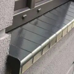 Porysowany parapet z blachy, z widocznymi przetarciami i drobnymi uszkodzeniami, stanowi element zewnętrzny budynku.