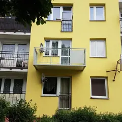 Żółty blok z dobudową balkonu na słupach. Widać okna, balustrady i anteny satelitarne.