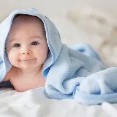 Ein lächelndes neugeborenes Baby liegt auf einem weißen Bett, eingewickelt in ein hellblaues Handtuch.