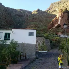 Un niño toma fotos en un camino empedrado, con una casa blanca y montañas rojas de fondo, cerca de la cueva bermeja.