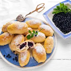 Pyszne jagodzianki posypane cukrem pudrem, obok miseczka świeżych jagód. Idealne na lato, zgodnie z najlepszym jagodzianki przepis.