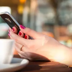 Dłoń z pomalowanymi paznokciami trzyma smartfon, obok biała filiżanka. Długą żywotność baterii w telefonie doceni każda użytkowniczka.