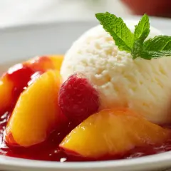 Ein klassischer Pfirsich Melba: Pfirsichspalten in rotem Beerensirup, garniert mit einer Kugel Vanilleeis, frischen Himbeeren und Minze.