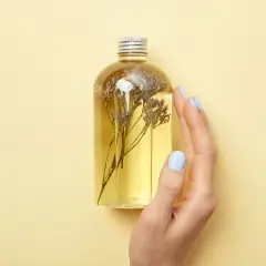 Eine Hand mit hellblauen Nägeln berührt eine Flasche mit gelbem Öl und getrockneten Blumen. Perfekt für alle, die Duschgel selber machen wollen.