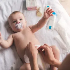 Chłopiec z smoczkiem leży na ręczniku, a ręce mamy dokonują **przewijania chłopca**. Obok kosmetyki i chusteczki.