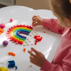 Dziecko tworzy tęczę z plasteliny, rozwijając kreatywne zabawy dla dzieci w domu. Kolorowe kuleczki i paski plasteliny tworzą magiczny świat.