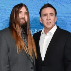 Weston Coppola Cage z długimi włosami i Nicolas Cage w czarnym garniturze pozują do zdjęcia na niebieskim tle.