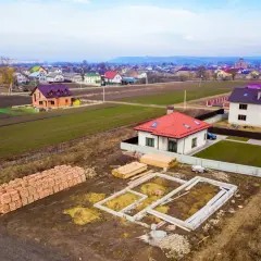 Budowa domku na działkę zrób to sam. Widać fundamenty, sterty cegieł i drewno budowlane. W tle ukończone domy.