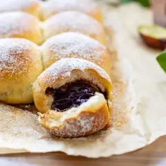 Puszyste ciasto krucho drożdżowe z dżemem śliwkowym, posypane cukrem pudrem. Obok połówki śliwek i zielone listki.