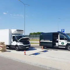 Kontrola drogowa: biała ciężarówka z otwartą maską i stosy palet obok, obok zielony bus Inspekcji Drogowej. Sprawdzana jest ładowność w dowodzie rejestracyjnym.
