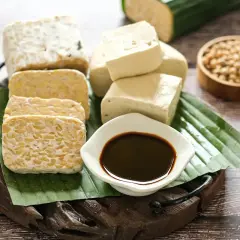 Tempeh und Tofu, zubereitet aus indonesischen Sojabohnen, liegen auf Bananenblättern. Dazu eine Schale dunkler Sojasauce und rohe Sojabohnen.
