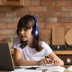 Dziewczynka w słuchawkach skupiona na nauczaniu domowym przy laptopie, z zeszytem i długopisem w dłoniach.