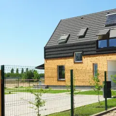 Nowoczesny pasywny dom modułowy z drewnianą elewacją i panelami słonecznymi na dachu, otoczony zielonym ogrodem i ogrodzeniem.