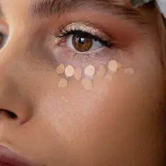Nahaufnahme einer Frau, die Concealer unter den Augen aufträgt. Mehrere helle Tupfer bilden eine Reihe, bereit zum Verblenden.