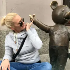 Kobieta w okularach przeciwsłonecznych i szarej bluzie bawi się z pomnikiem misia, który trzyma tabliczkę. To część szlaku łodzi bajkowej.