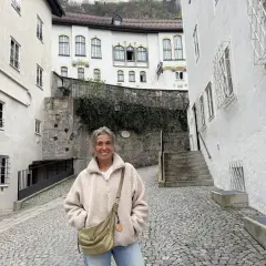 Eine Frau steht lächelnd auf einem Kopfsteinpflasterweg zwischen alten weißen Häusern. Sie könnte eine Pilgerin sein, die auf ihrer Reise neue Orte entdeckt.