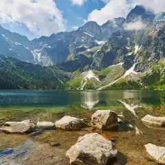 Widok na Morskie Oko z kamienistym brzegiem. W tle majestatyczne góry i lasy. Palenica Białczańska w oddali.