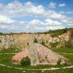 Krajobraz po kamieniołomie w Wietrzni w Kielcach. Skarpy, hałda ziemi i zieleń pod błękitnym niebem z chmurami.