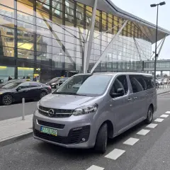 Szary van czeka na pasażerów przed nowoczesnym terminalem lotniska. Może to być transport z Koszalina do Gdańska.