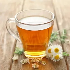 Ein Glas mit goldenem Kamillentee steht auf Holz. Kleine Kamillenblüten schmücken die Szene. Perfekt, um kamillentee kalt trinken.