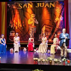 En el teatro Los Corrales de Buelna, cinco damas de honor y un presentador esperan en el escenario bajo un telón de San Juan.