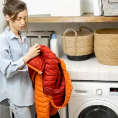 Kobieta trzyma pomarańczową kurtkę, przygotowując się do prania. W tle pralka i kosze.