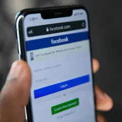 Dłoń trzyma smartfon z ekranem logowania Facebooka. Widoczny formularz do wpisania numeru telefonu/emaila i hasła, przycisk "Log In" oraz opcja "Forgot Password" - pomoc w sytuacji, jak odzyskać konto na FB.