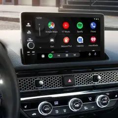 Wnętrze samochodu z ekranem Android Auto, na którym widać ikony Google Maps, Spotify i innych aplikacji. W tle dyskretnie widać Yanosik.