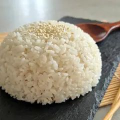 Ein Berg aus weißem Reis, bestreut mit Sesam, auf einem schwarzen Stein. Daneben liegen Stäbchen und ein Holzlöffel. Das ist die Basis, was bedeutet Sushi.
