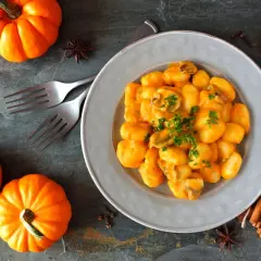 Pyszne gnocchi z kremowym sosem dyniowym, ozdobione natką pietruszki. Obok talerza małe dynie i przyprawy.
