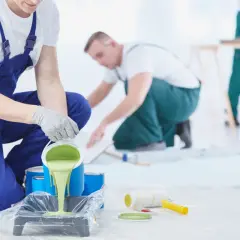 Målare häller ljusgrön färg i en tråg för att måla furugolv. Verktyg som roller och pensel ligger redo.