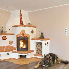 Ein Hund liegt vor einem großen, weißen Kachelofen mit integriertem Kamin. Der Ofen hat eine Sitzbank und ist mit bunten Kacheln verziert. Der Unterschied zwischen Kamin und Ofen wird hier durch die massive Bauweise und die Heizfunktion des Ofens deutl...