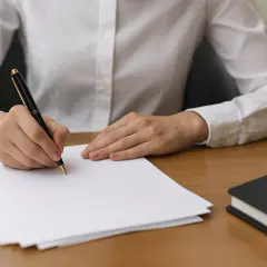 Main d'une personne écrivant sur du papier avec un stylo, à côté d'un carnet.